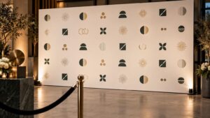 Warmly lit event entrance with a large cream step-and-repeat backdrop printed with repeating dark green and gold abstract geometric marks, framed by plants, candles, velvet rope stanchions, and stone pedestals.